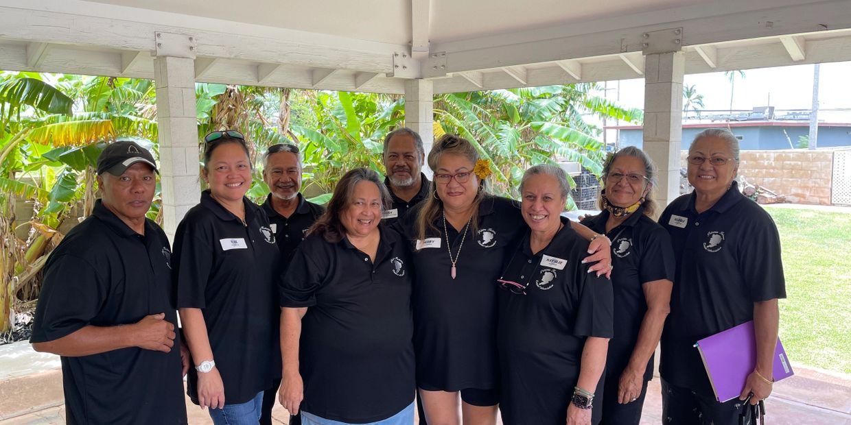 Nine members of the Princess Kahanu Estates Association standing together in matching black polo shirts under a community pavilion.